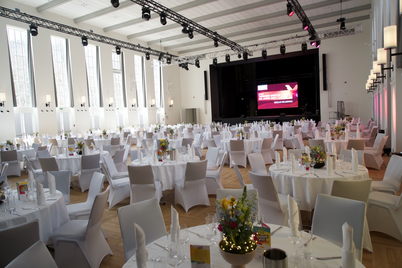 Heller Saal der Festhalle Ilmenau mit Rundbogenfenstern, runden Tafeln, Blumen und Bühne mit Leinwand