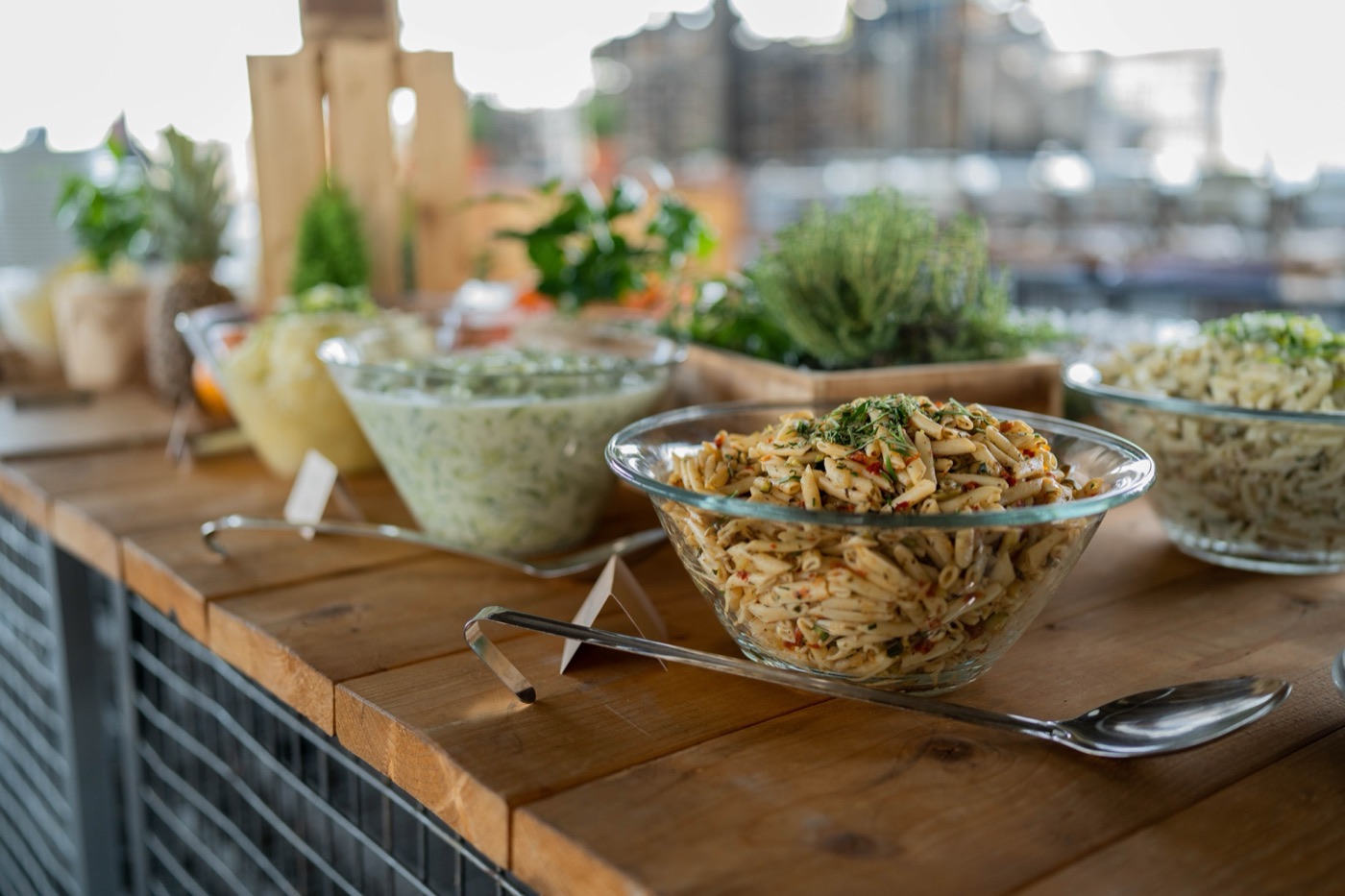 Buffet mit Nudelsalaten, Kräutern und Servierlöffeln auf einer Holztheke