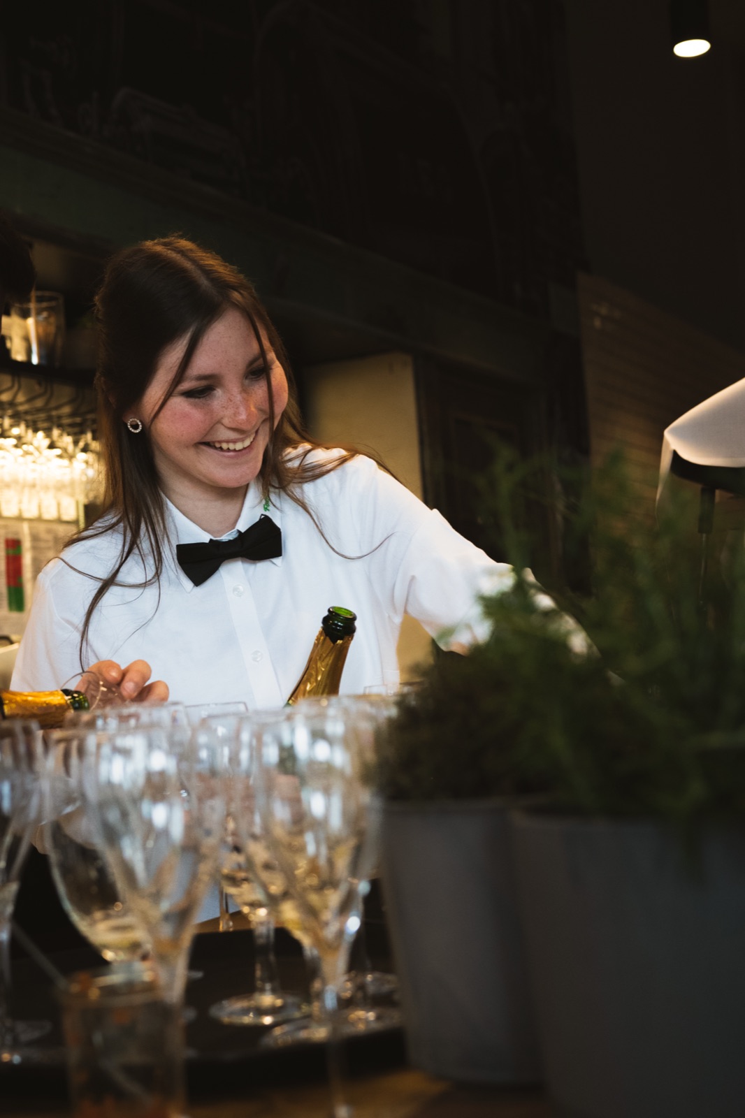 Eine Servicekraft im weißen Hemd mit schwarzer Fliege hinter einer Bar mit vorbereiteten Gläsern