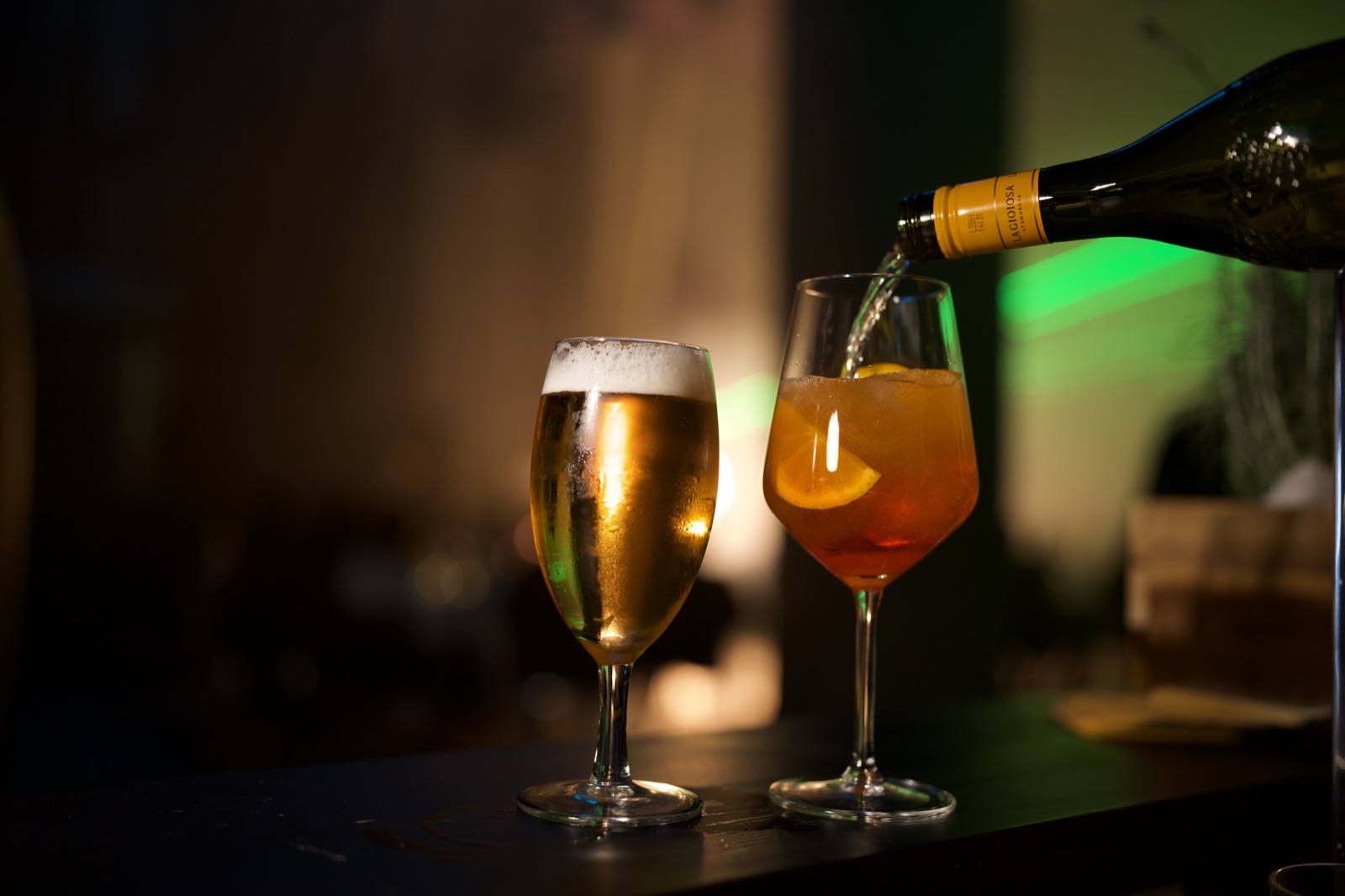 Ein Spritz mit Orangenscheibe wird aufgegossen; daneben steht ein gefülltes Bierglas